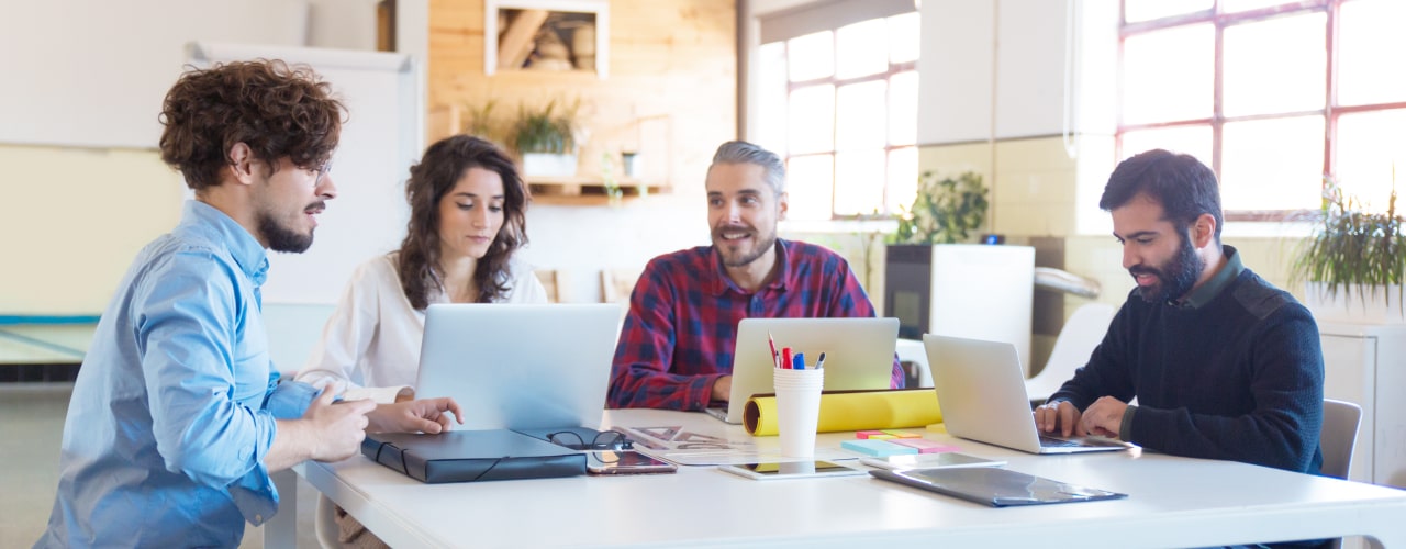 Hero image featuring a diverse team of professionals in a work setting, engaged in a meeting and working on laptops.